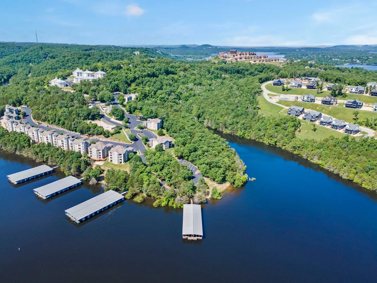 A scenic view of the lakefront with residential buildings and boathouses nestled among lush greenery. The calm water reflects the sky, while the setting hints at private and communal spaces for both relaxation and adventure. The ambiance is serene yet inviting for various outdoor activities.