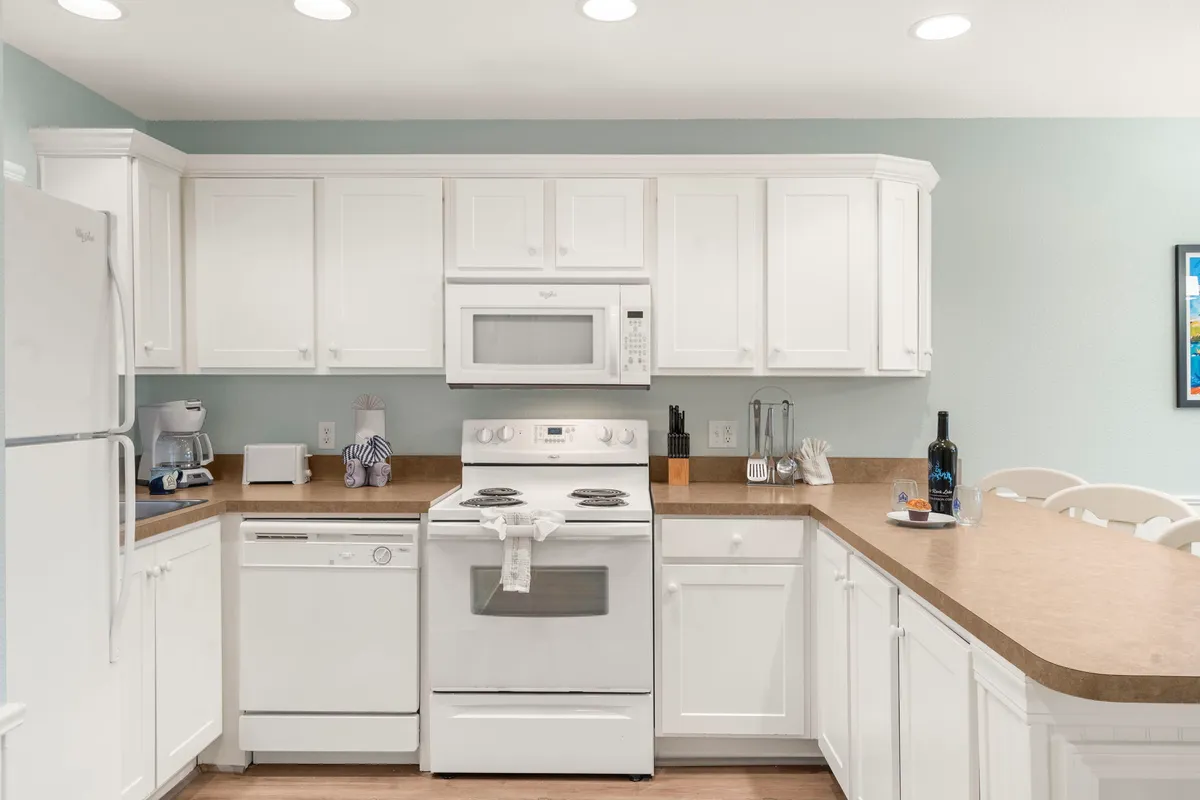 A bright, cozy kitchen space showcasing white cabinetry, stainless steel appliances, and a central kitchen island. The scene radiates warmth and comfort, inviting guests to cook, bake, or simply gather around. The design blends practicality with a welcoming ambiance.