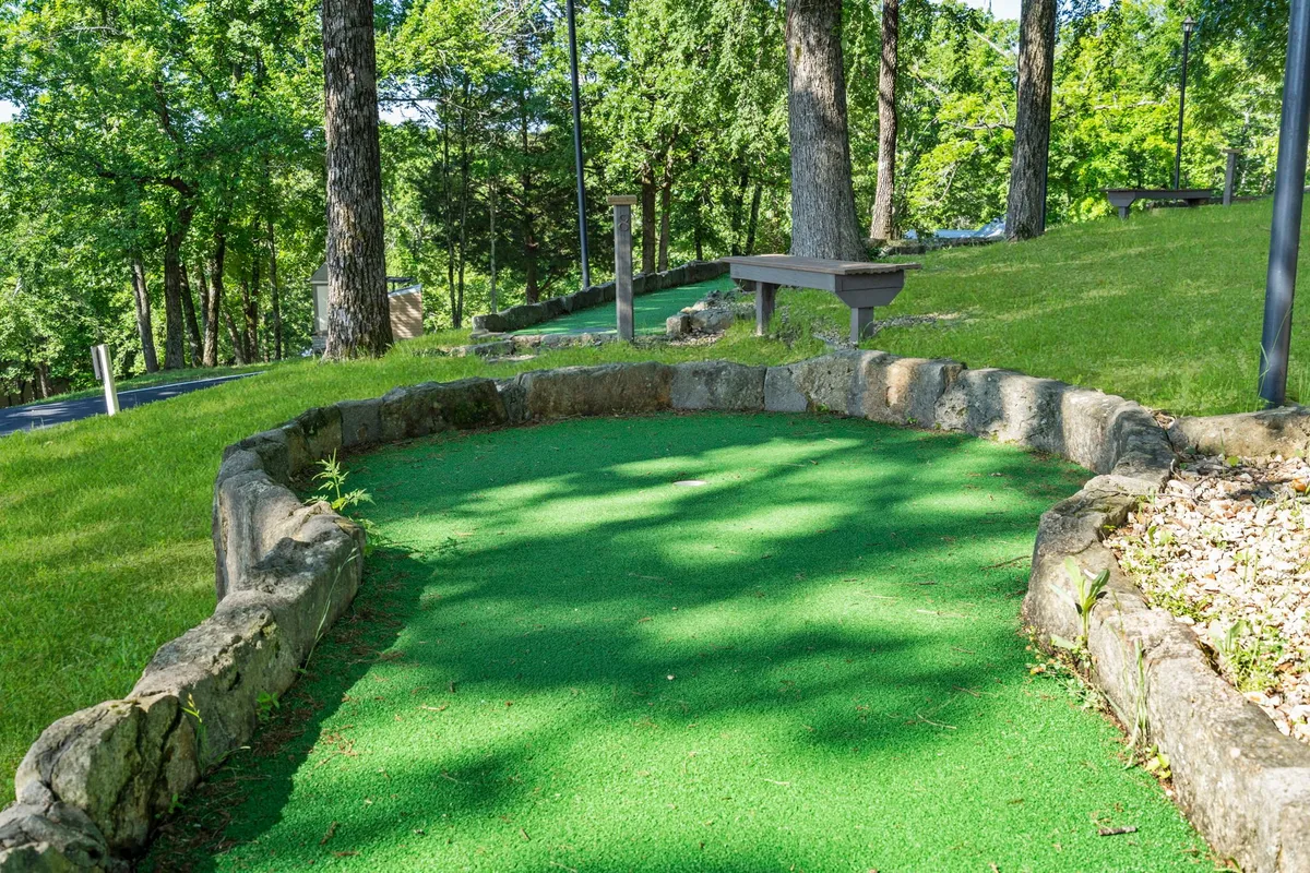 A close-up of a mini-golf green bordered by stone, nestled among mature trees with benches nearby. The playful environment is ideal for family competitions and casual relaxation in nature.