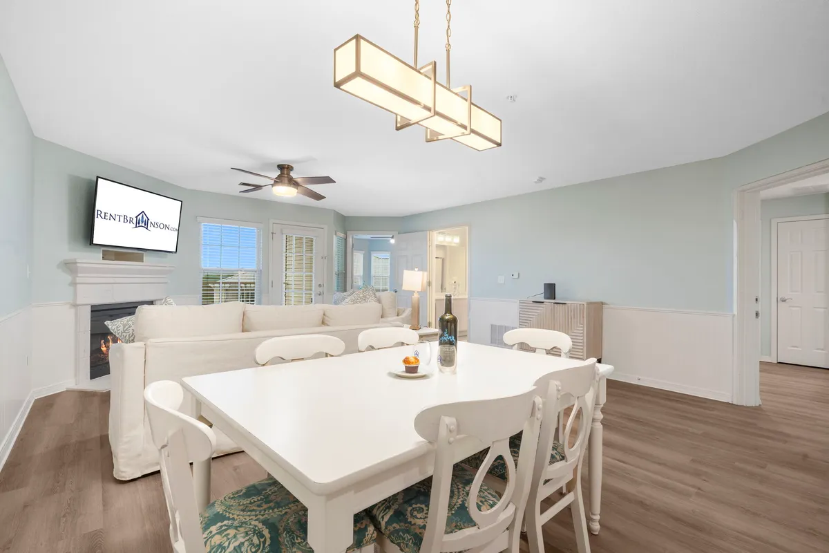 An open-plan space featuring a dining table set for a meal, a comfortable sofa, a mounted television, and a stylish light fixture. The sofa and dining area flow together, emphasizing an open, communal environment.