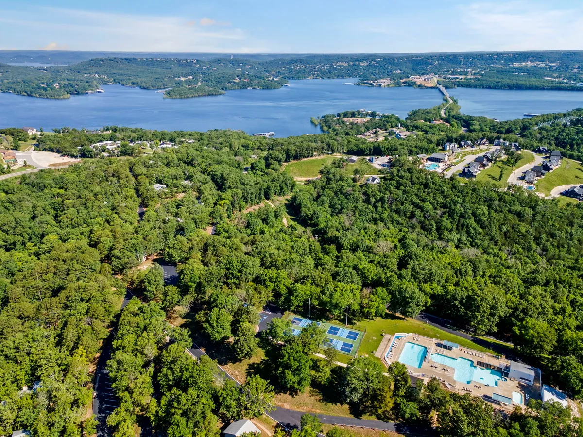 An expansive aerial view showing a large lake bordered by lush greenery, with a prominent swimming pool area and tennis courts in the foreground. The image captures the scale of the property, its natural surroundings, and highlights resort-style amenities such as the pool and courts.