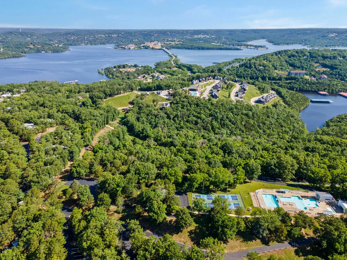 A sweeping view of the property showing the lake, surrounding greenery, outdoor swimming pool, tennis courts, and residential buildings. The scene exudes a relaxed, social, and recreational mood, highlighting a variety of resort activities within walking distance.