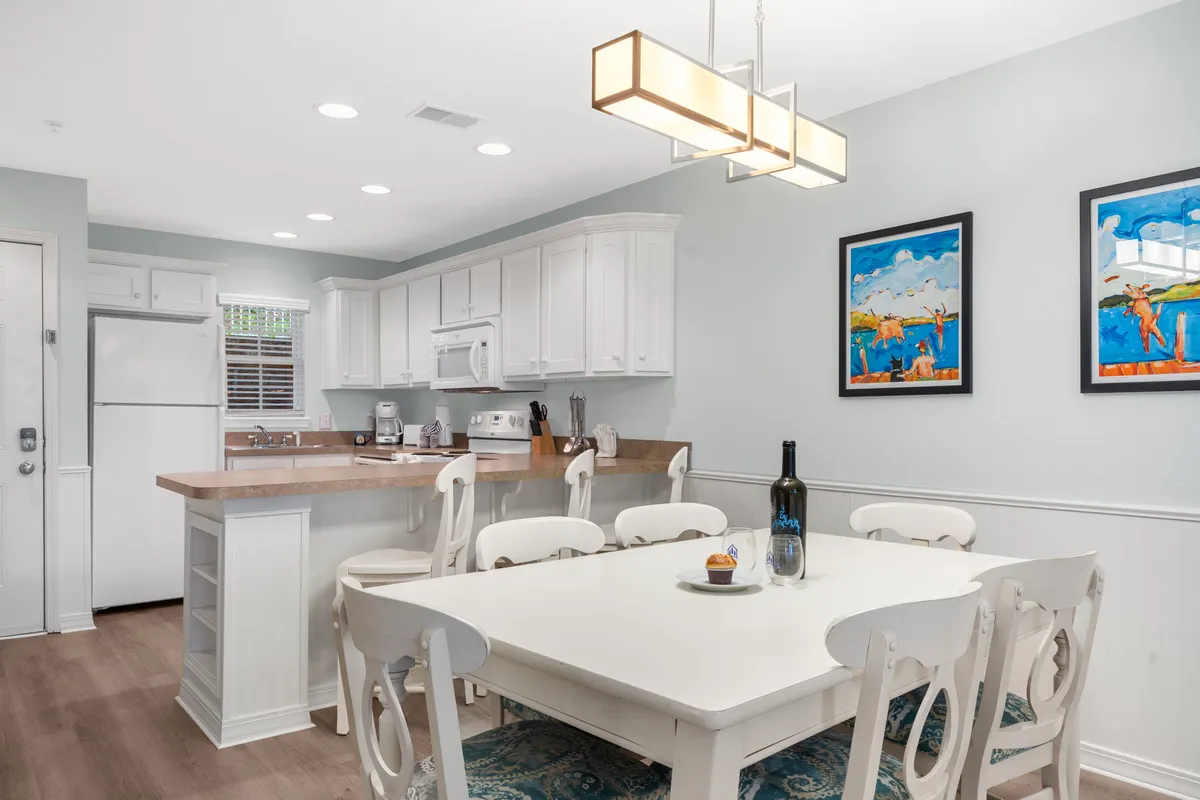A dining area featuring a large table set beneath a sparkling chandelier, with a view into the kitchen countertop and tasteful wall art. The setup suggests a space ready for hearty family meals, celebrations, or lively board game nights. The ambiance is warm, welcoming, and ideal for group gatherings.