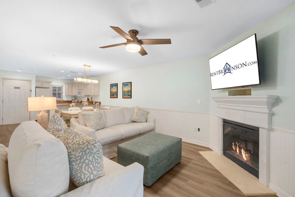 A cozy living room with a comfortable sofa arranged beside a fireplace, a television above the mantel, and tasteful decorative artwork on the walls. The space is well lit, inviting, and clearly designed for relaxation and gathering.