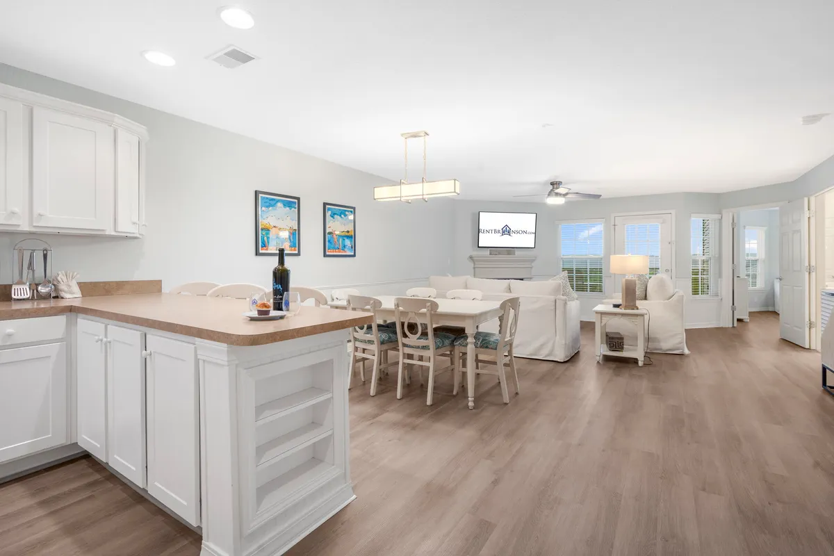 A bright interior shot showing a kitchen island, dining table, and living area seating, with artwork adorning the walls. Large windows let in ample natural light and offer tranquil outside views, further enhancing the inviting mood.