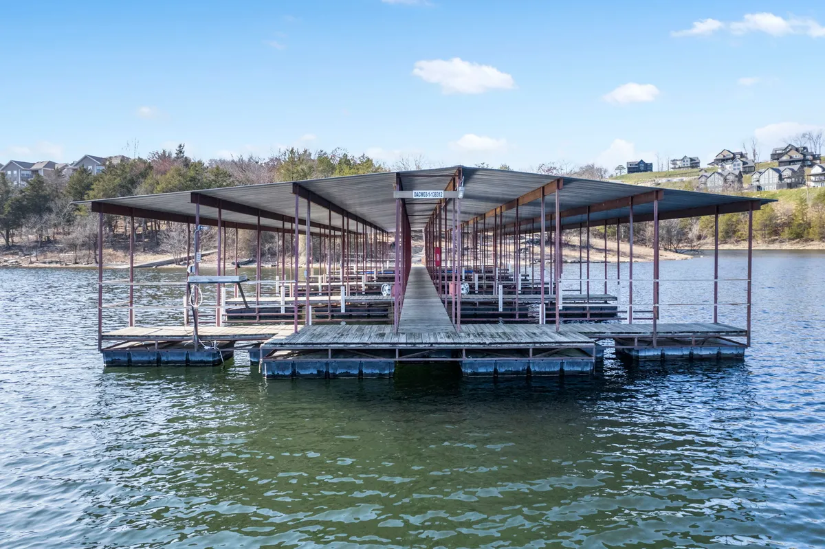 A wooden dock extends over calm lake waters, framed by the surrounding landscape of trees and gentle hills. The peaceful scene is ideal for fishing, relaxing, or preparing to set off in a boat. The expansive water view emphasizes the direct connection between the property and the lake.
