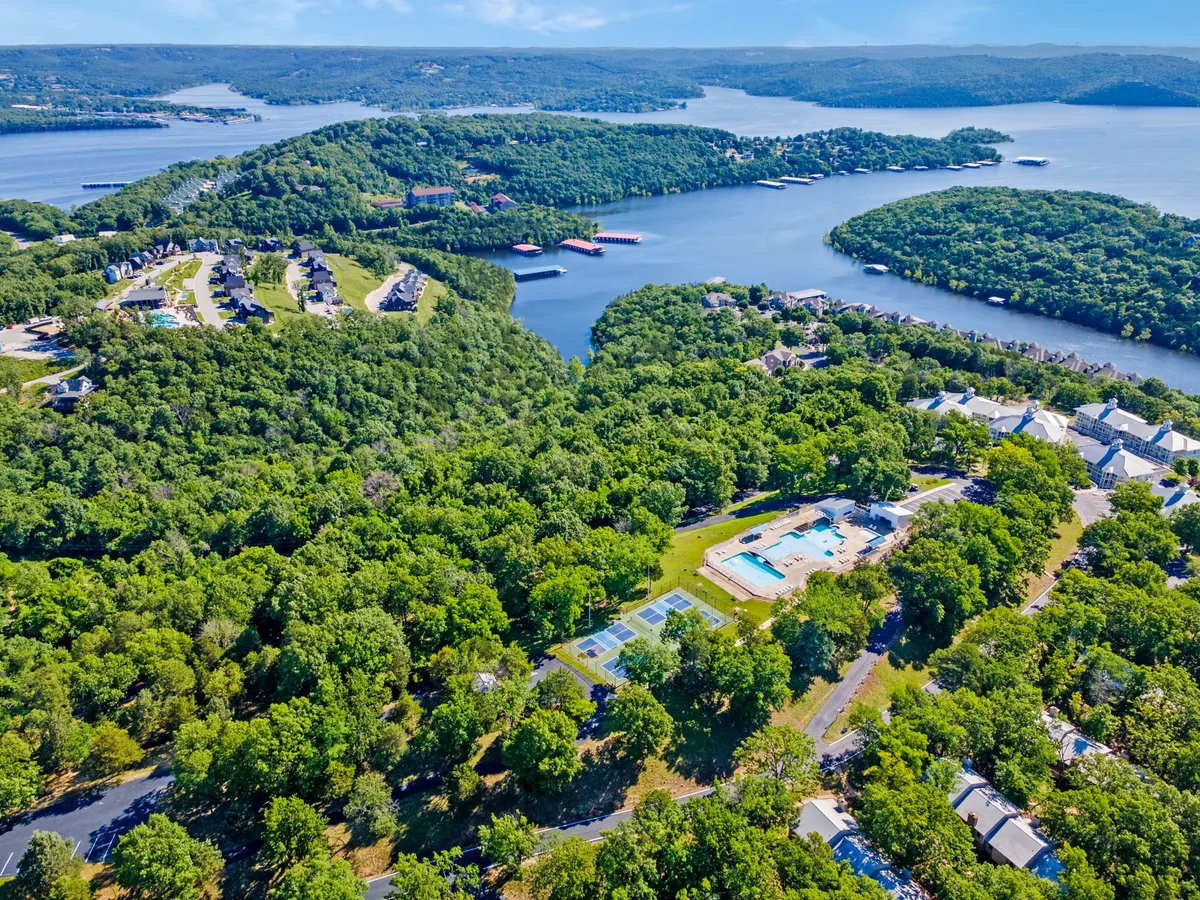 A wide exterior shot that encompasses a swimming pool, tennis courts, lush green trees, Table Rock Lake, and scenic hills. This view highlights a range of resort amenities and the beautiful lake setting, with opportunities for swimming, tennis, boating, hiking, and picnicking.